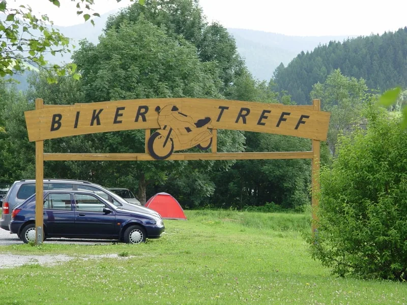 Holzschild „Biker Treff“ über einer Wiese in Rettenegg, dahinter Bäume und Hügel.