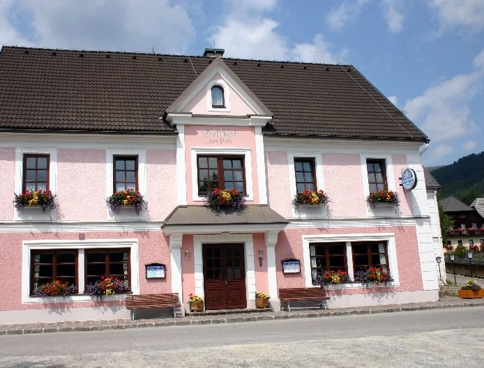 Das rosa Haus des Gasthofs zur Post in Rettenegg mit weißen Fensterfaschen, Blumenkästen voller Geranien und Holzbänken vor dem Eingang.