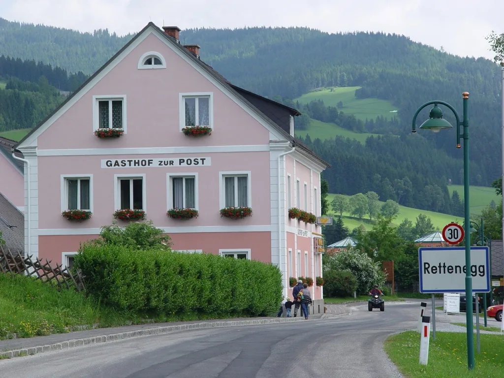 Der rosa Gasthof zur Post an der Straße in Rettenegg, davor die Ortstafel „Rettenegg“, dahinter bewaldete Hügel.
