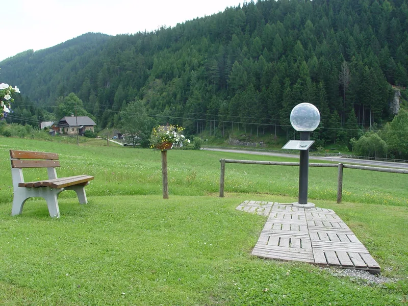 Station des Planetenwegs: eine Bank und eine Planetenkugel auf einer Wiese, dahinter Wald.